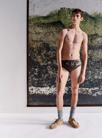 A shirtless young man in dark briefs, grey socks, and tan shoes stands indoors in front of a mottled earth-tone background poster.