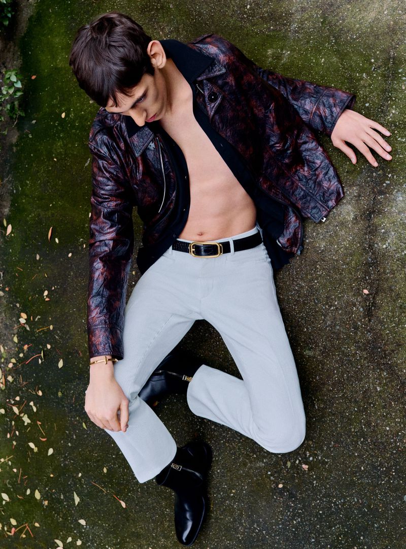 Overhead view of a young adult male fashion model with dark hair, wearing a printed leather jacket and light denim jeans.