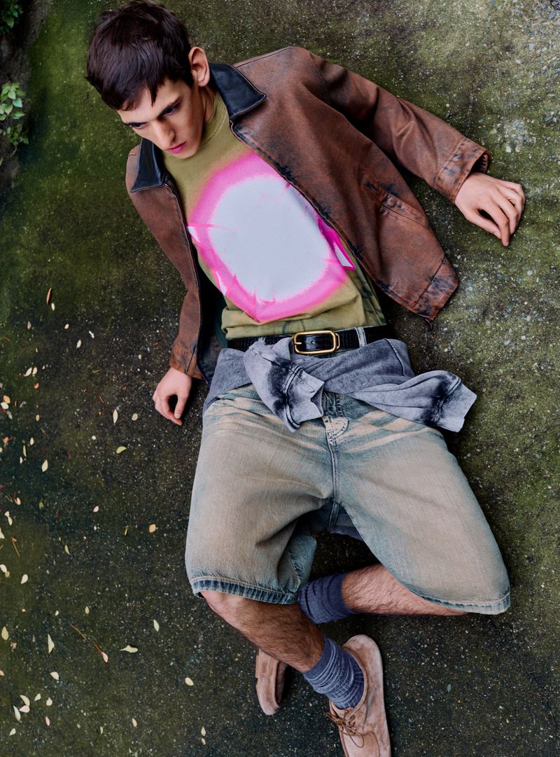 Male model wearing a brown leather jacket and patterned t-shirt lying on a mossy ground for Le Mile Magazine.