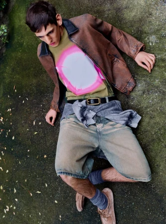 Male model wearing a brown leather jacket and patterned t-shirt lying on a mossy ground for Le Mile Magazine.