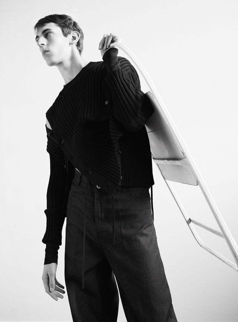 Black and white fashion shot of a slender young adult wearing a knit cardigan and dark jeans while holding a white folding chair.