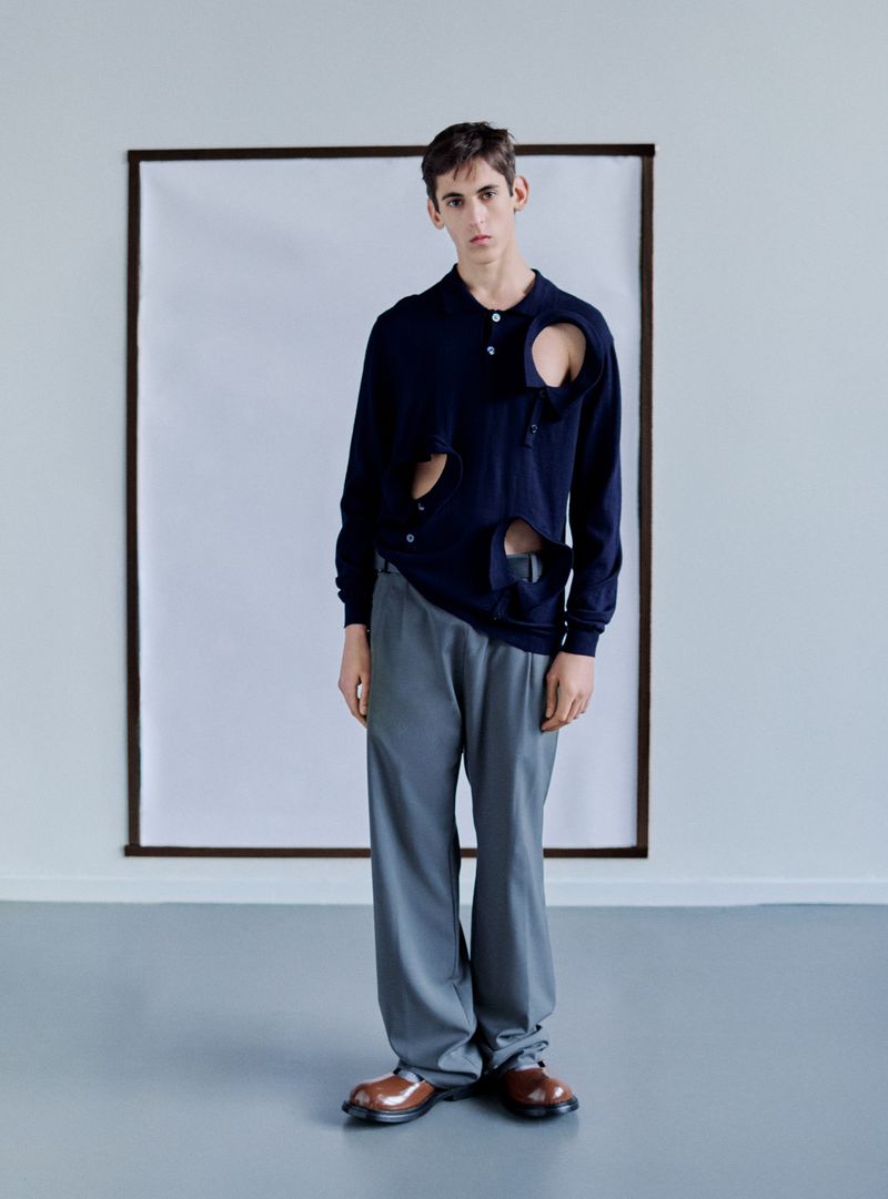 A male model poses in a navy long-sleeve shirt with irregular cutouts, paired with grey trousers and brown leather shoes, against a white backdrop.