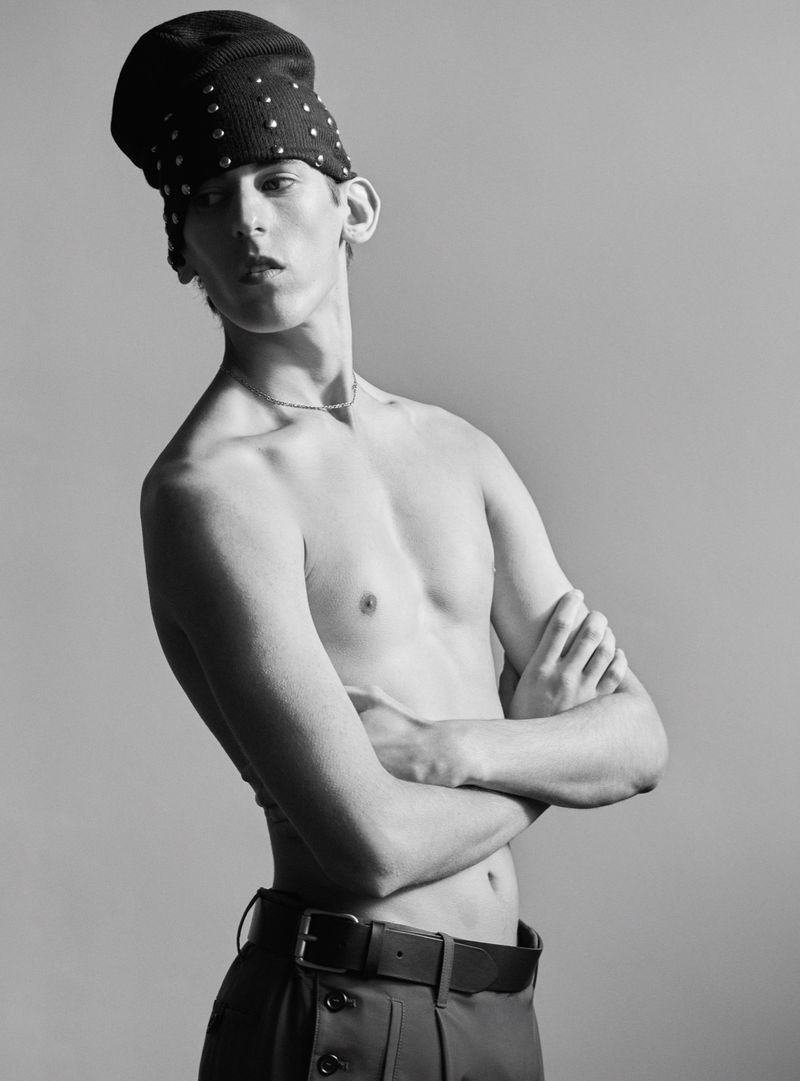 Monochrome fashion portrait of a shirtless male model wearing a studded cap against a grey studio background