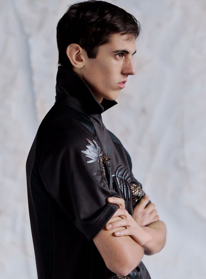 A young adult man in a black short-sleeved shirt with decorative plant designs poses with arms crossed in a studio with a white backdrop.