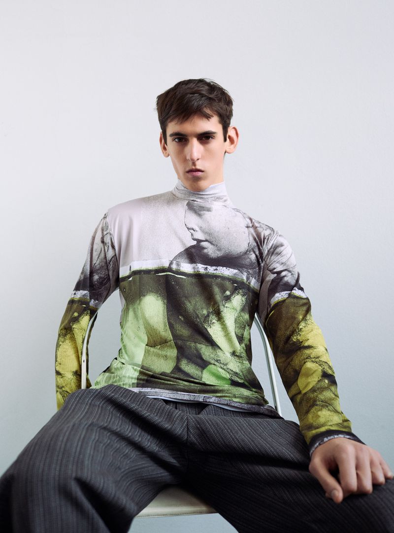 Male model seated in a chair wearing a graphic print turtleneck and dark trousers against a white wall.
