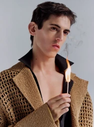 Close-up portrait of a male model holding a lit match wearing a textured camel cardigan and black shirt against a grey background.