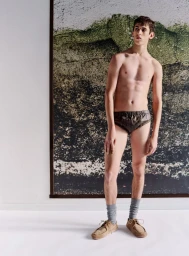 Shirtless male model wearing dark underwear, grey socks, and tan shoes standing before a large landscape artwork.