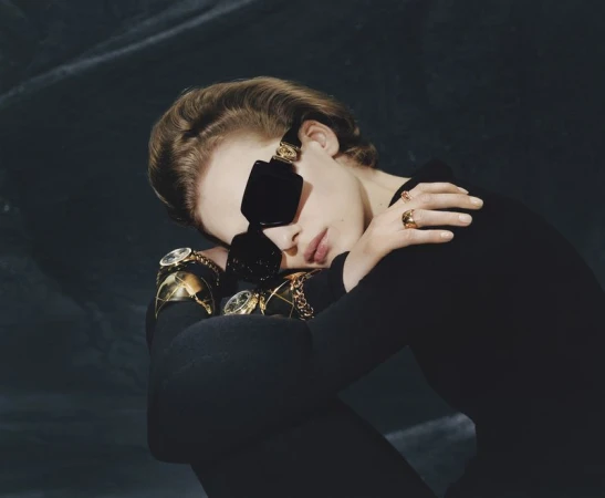 Model in black top wearing dark sunglasses and gold jewelry posing against a dark textured background