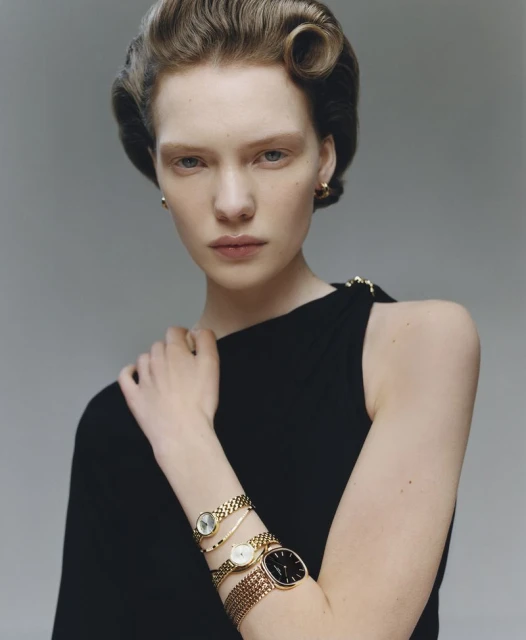 Close-up of a fair-skinned young woman with vintage wavy hair wearing a black dress and gold wristwatches in a studio setting.