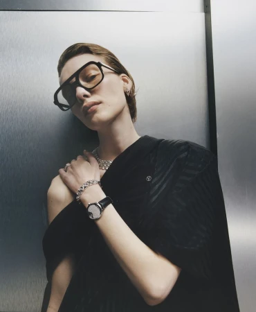 Model in oversized sunglasses and black top wearing diamond jewellery and a watch against a metallic background