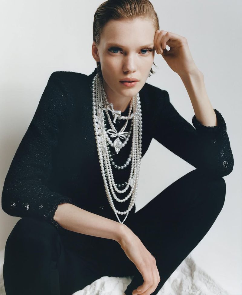 Woman with slicked-back hair poses indoors wearing a black jacket and multiple strands of pearl necklaces with pendant details.