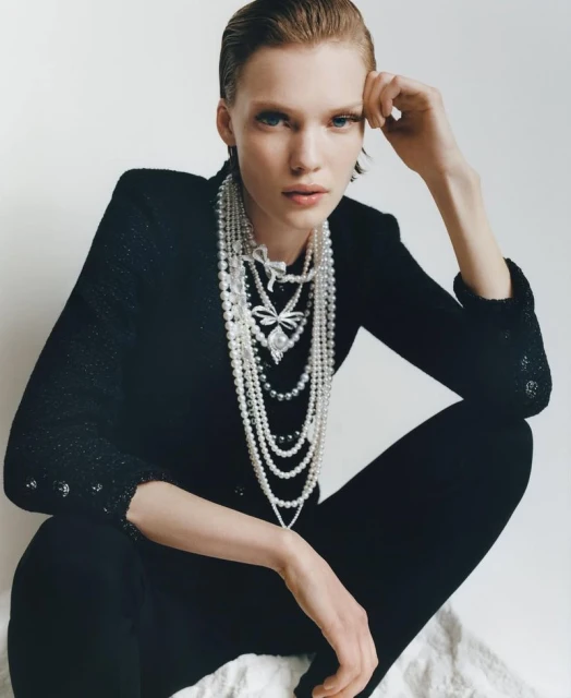 Woman with slicked-back hair poses indoors wearing a black jacket and multiple strands of pearl necklaces with pendant details.