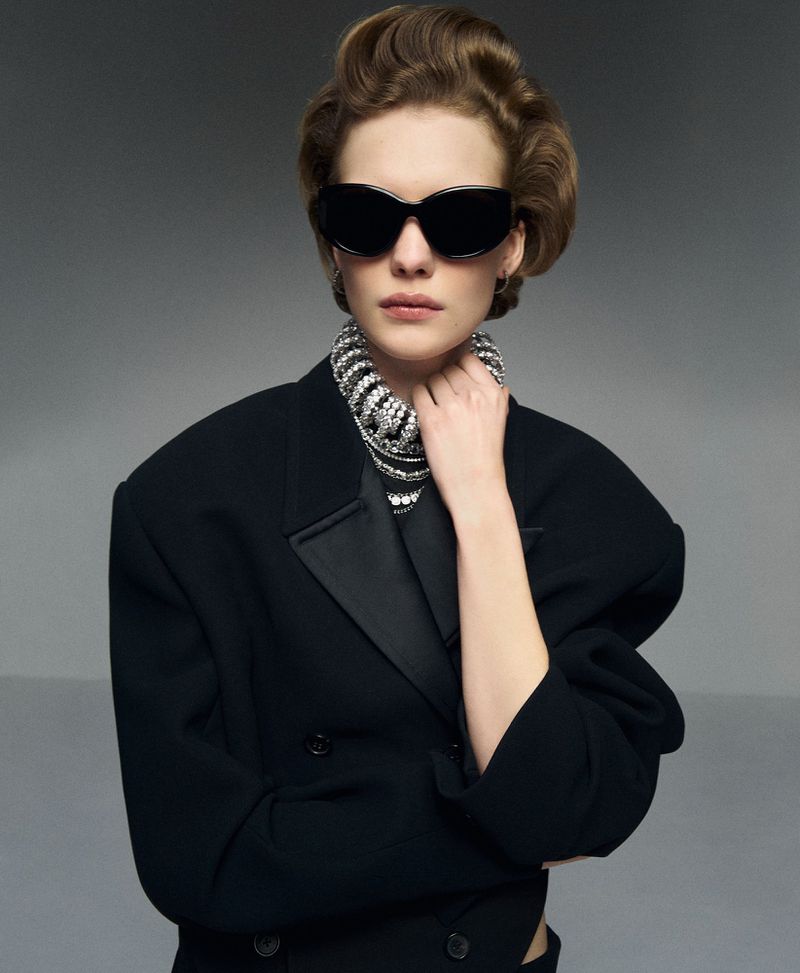 Model wearing black sunglasses, a statement rhinestone necklace, and an oversized black blazer against a grey backdrop.