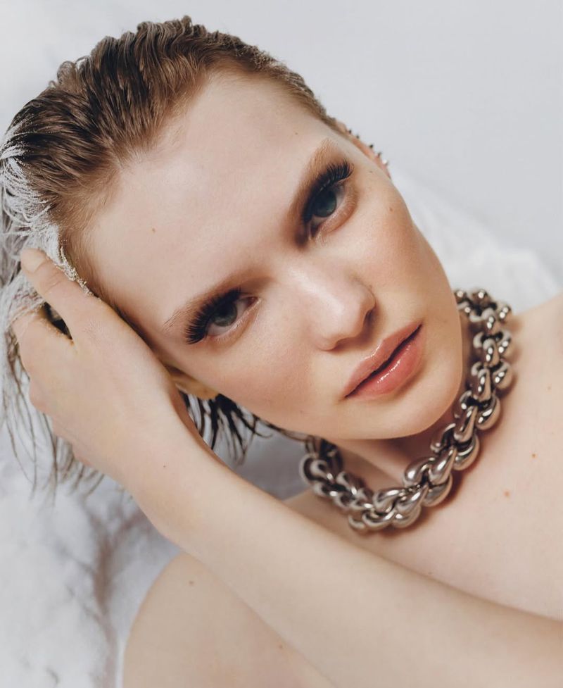 Close-up portrait of a young adult female model with silver chain necklace, wet hair, and light makeup against a white background.