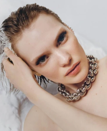 Close-up portrait of a young adult female model with silver chain necklace, wet hair, and light makeup against a white background.