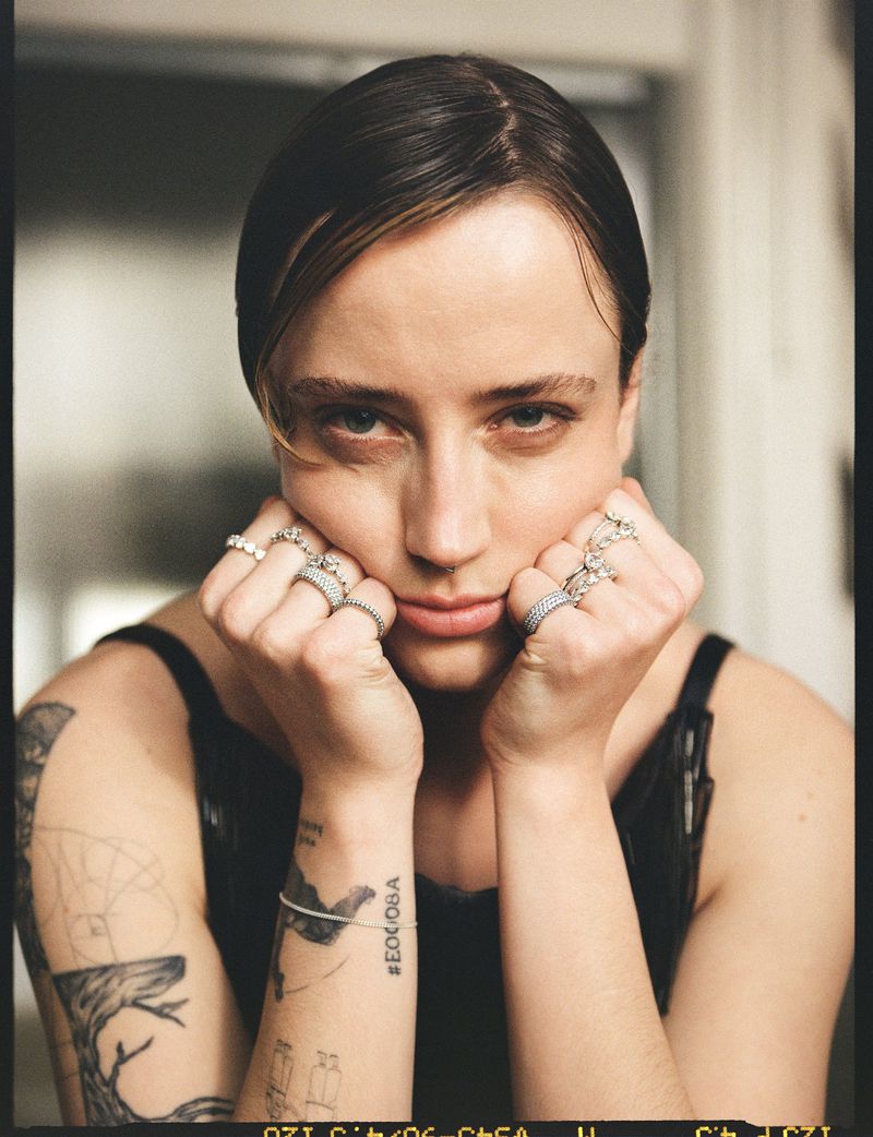 Close-up portrait of a woman resting her chin on her hands with rings on her fingers and visible arm tattoos against an interior background.