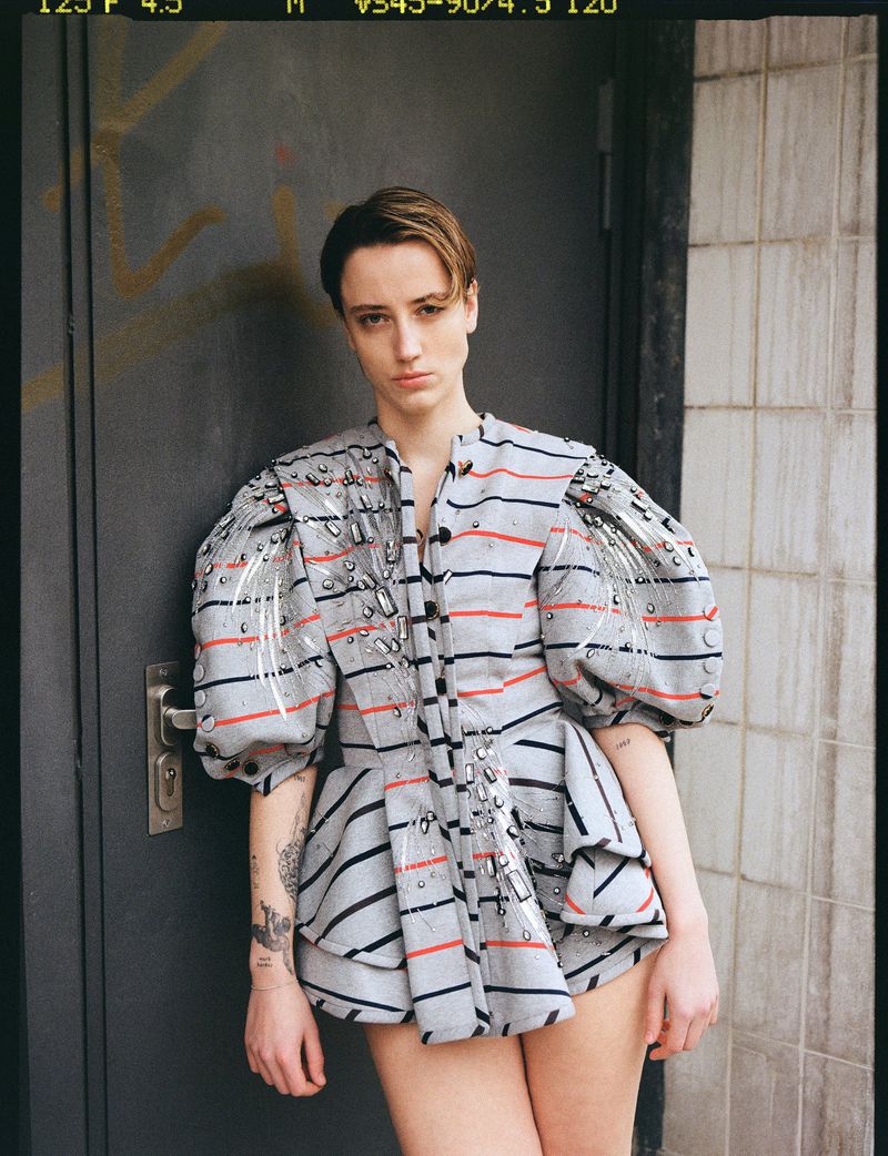 Portrait of a woman wearing a striped grey top with puffed sleeves, posing in front of a grey weathered door with graffiti.