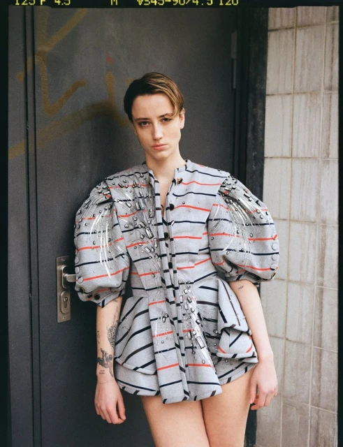Portrait of a woman wearing a striped grey top with puffed sleeves, posing in front of a grey weathered door with graffiti.