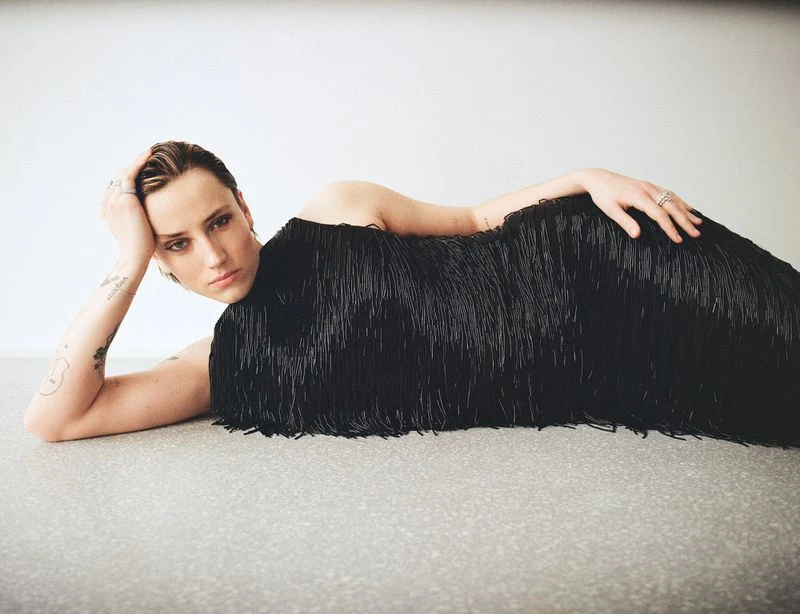 Person lying down in a black fringed dress on a gray floor, leaning on their left hand, looking at the camera in a studio setting.