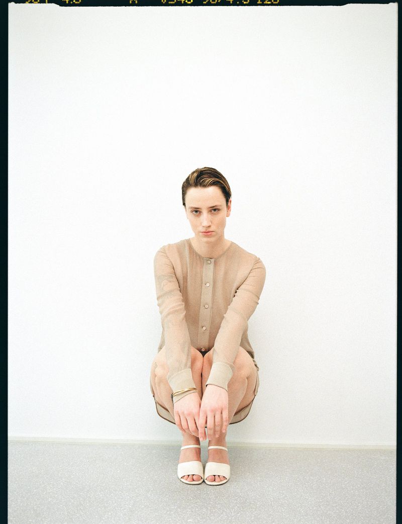 Portrait of a young woman in a sheer beige top and sandals, crouching against a white wall in a photo studio with soft, neutral lighting.