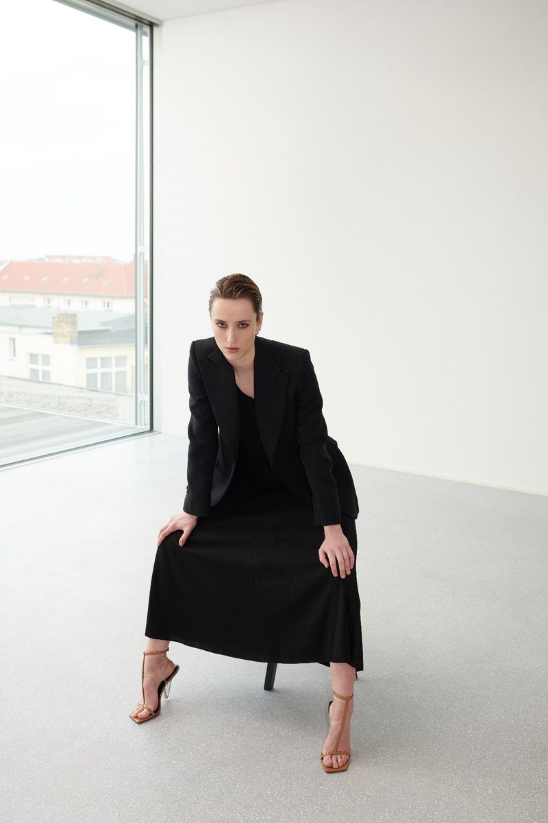 Woman seated wearing a black blazer and skirt with strappy heels in a minimalist white interior space with a tall window