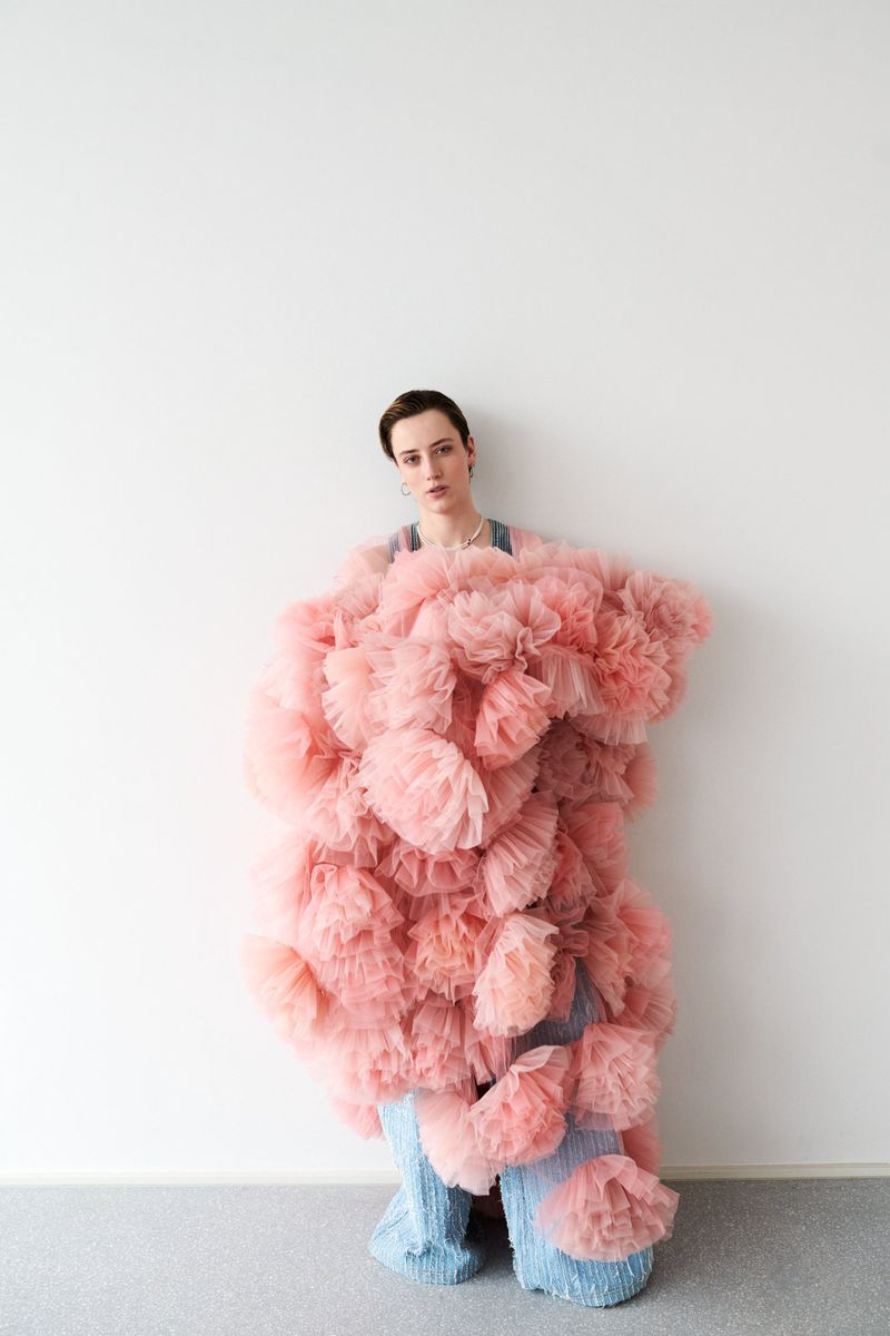 Woman in pink ruffled garment and wide-leg blue pants poses against a white wall in a fashion-style portrait in a studio setting.