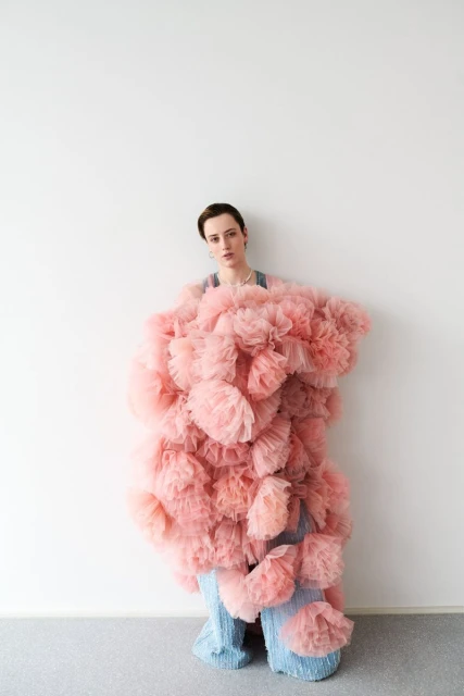 Woman in pink ruffled garment and wide-leg blue pants poses against a white wall in a fashion-style portrait in a studio setting.