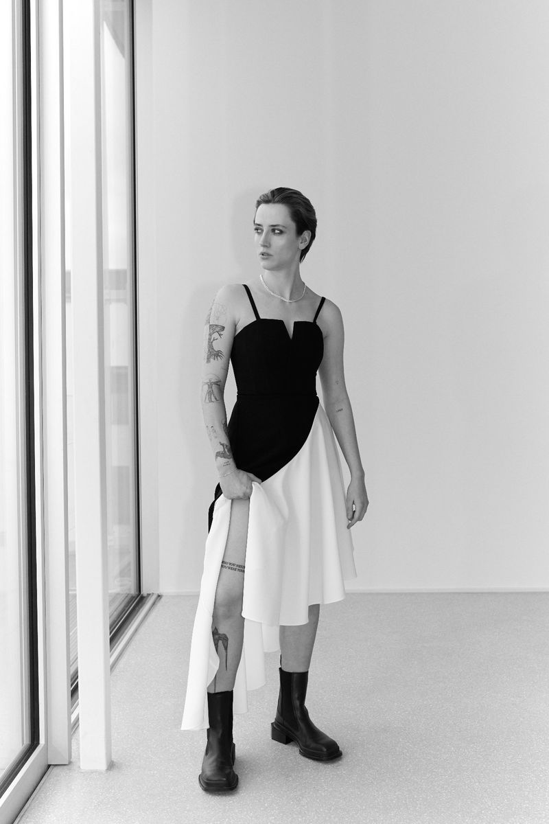 Monochrome portrait of a woman in a black and white dress and black boots posing inside a room next to a window with natural light.