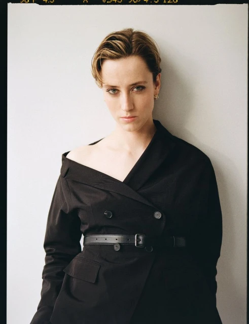 Portrait of a woman with short blonde hair wearing a black jacket with a belt against a plain white background in a studio setting.