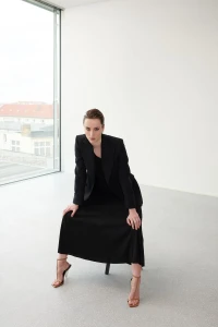 Woman seated wearing a black blazer and skirt with strappy heels in a minimalist white interior space with a tall window