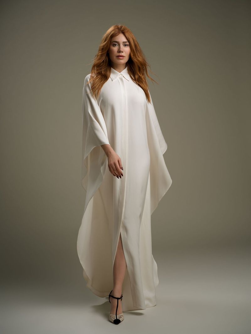Elegant redhead woman in a flowing white dress walks in a studio. Fashion portrait with soft lighting and a clean background.