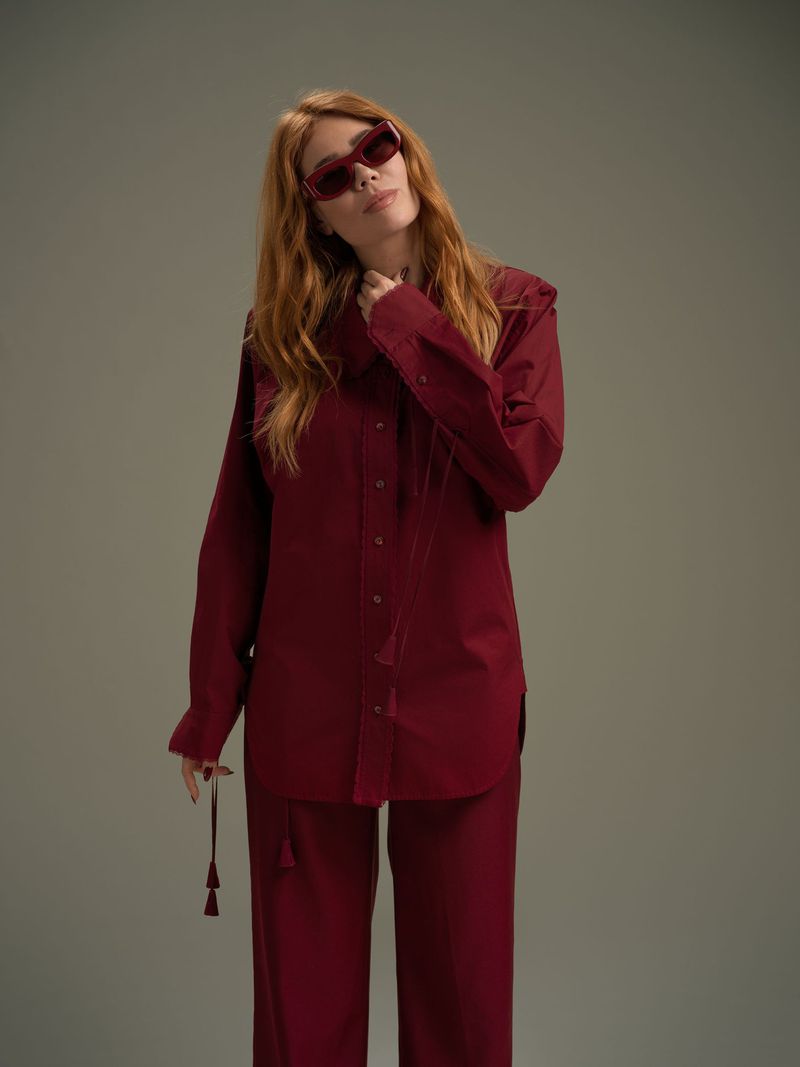 Portrait of a red-haired woman in a matching maroon pantsuit and sunglasses, posing indoors against a neutral backdrop with soft studio lighting.