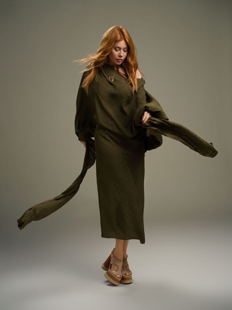 Red-haired woman in a green dress stands in a studio, looking down with arms spread and hair flying, showcasing a fashion-forward style.