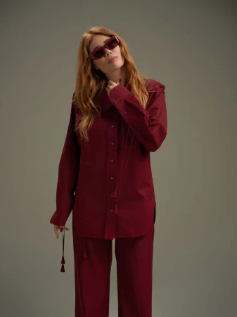 Palina Rojinski wearing a burgundy button-down shirt and matching trousers with dark sunglasses against a plain grey backdrop.