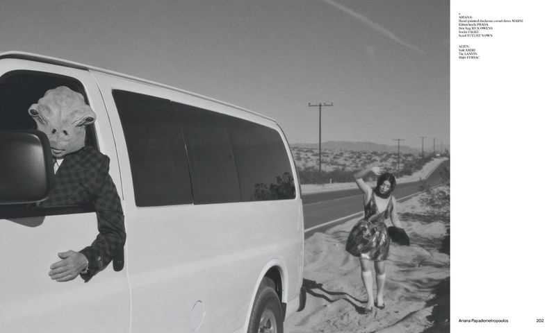 An alien in a suit leans out of a van window on a desert road, a woman waves nearby, monochrome desert landscape and sky.