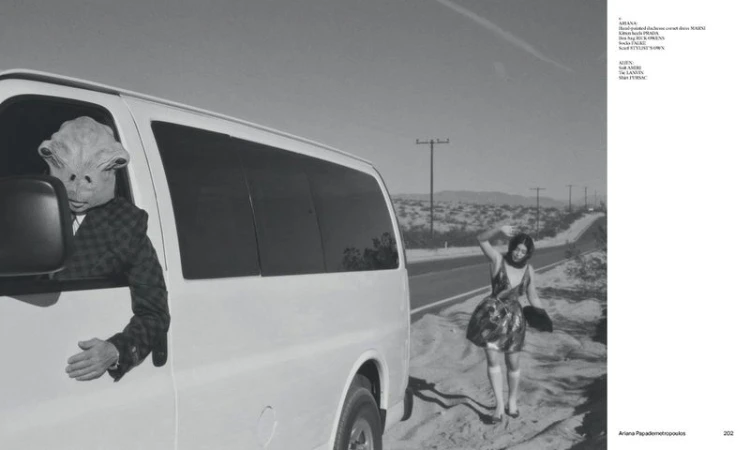An alien in a suit leans out of a van window on a desert road, a woman waves nearby, monochrome desert landscape and sky.