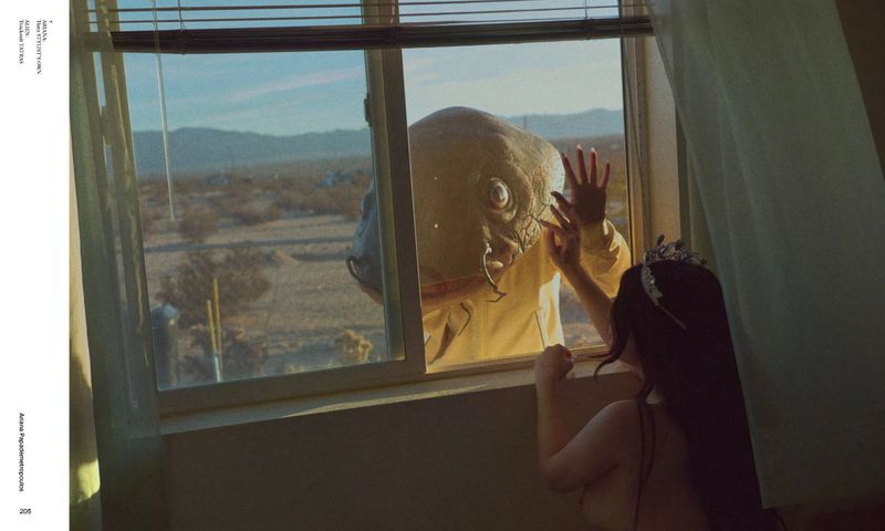 Woman in a tiara looks out a window at a figure wearing a reptile mask in a desert landscape, creating a surreal, unsettling image.