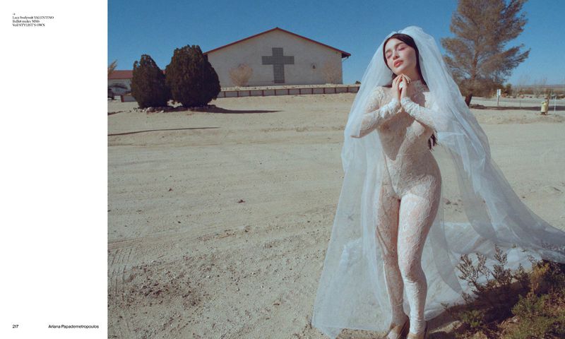 Woman in a lace jumpsuit and veil poses in a desert landscape with a building featuring a cross, beneath a clear blue sky.
