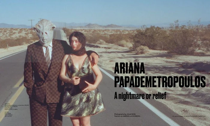 A woman in a metallic dress and a person in a plaid suit wearing an alien mask stand on an empty desert road.