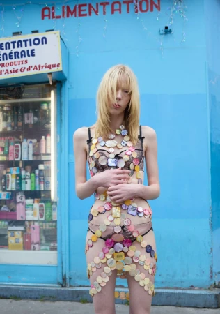 Adult female posing outdoors in decorative lingerie made of bottlecaps, standing in front of a blue convenience store storefront.