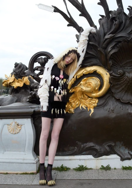 A fair-skinned, blonde model in a fur-trimmed jacket poses outdoors, leaning against a black and gold decorative wall with an overcast sky above.