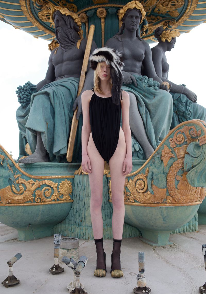 Fashionable woman poses in a black swimsuit and a fur hat in front of an ornate water fountain with statues in an outdoor setting.