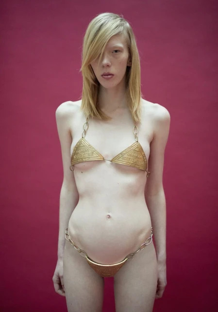 A slender blonde woman poses against a solid pink background, wearing a unique gold-colored bikini-like outfit made of chains and metal pieces.