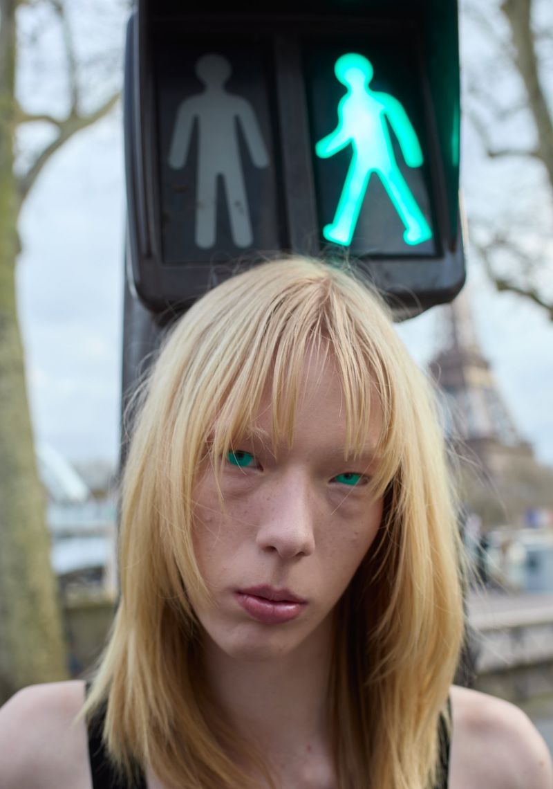 Portrait of an androgynous adult with blonde hair and light eyes under a pedestrian crossing signal light with the Eiffel Tower in the back.
