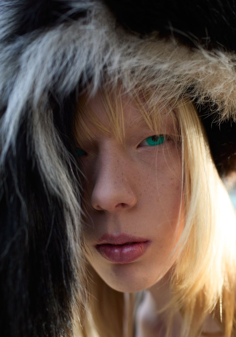 Close-up portrait of a fair-skinned adult wearing a fur-trimmed hat, showcasing a distinctive beauty look with pale blonde hair and striking green eyes.