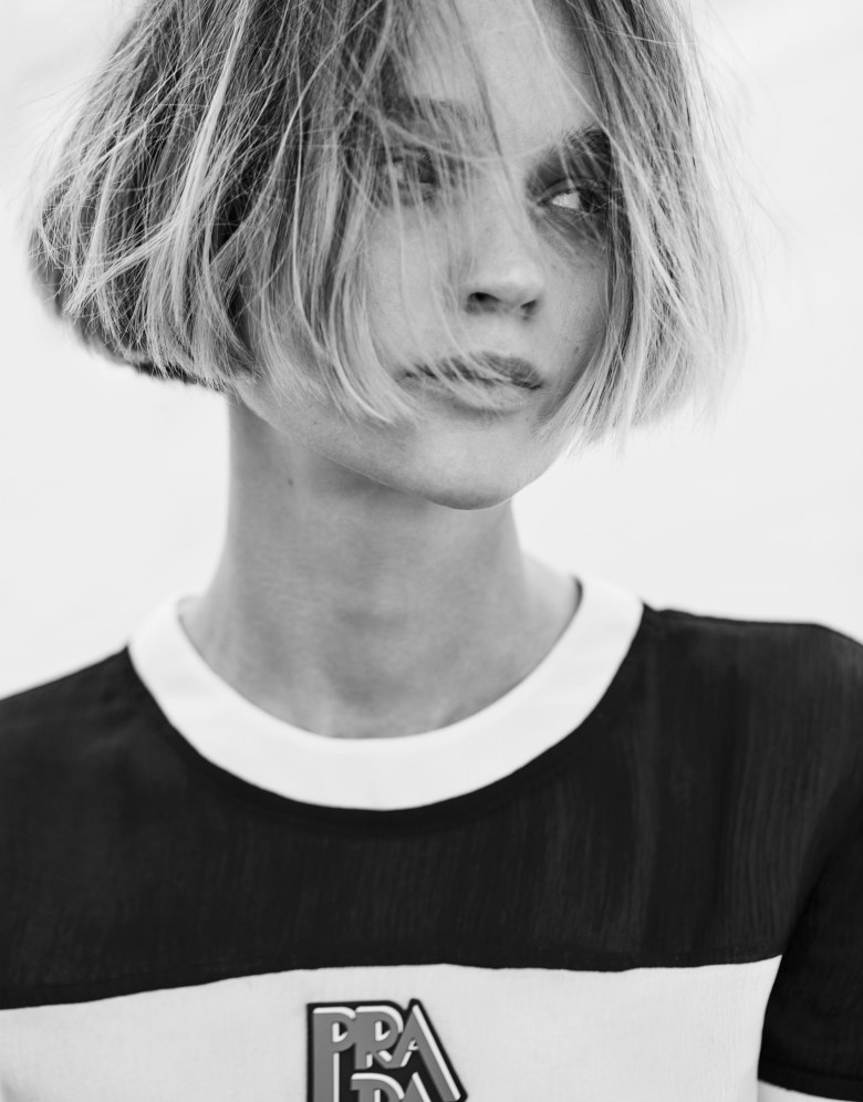 Black and white portrait of an adult female with a short bob haircut, wearing a shirt with a Prada logo, against a bright background.
