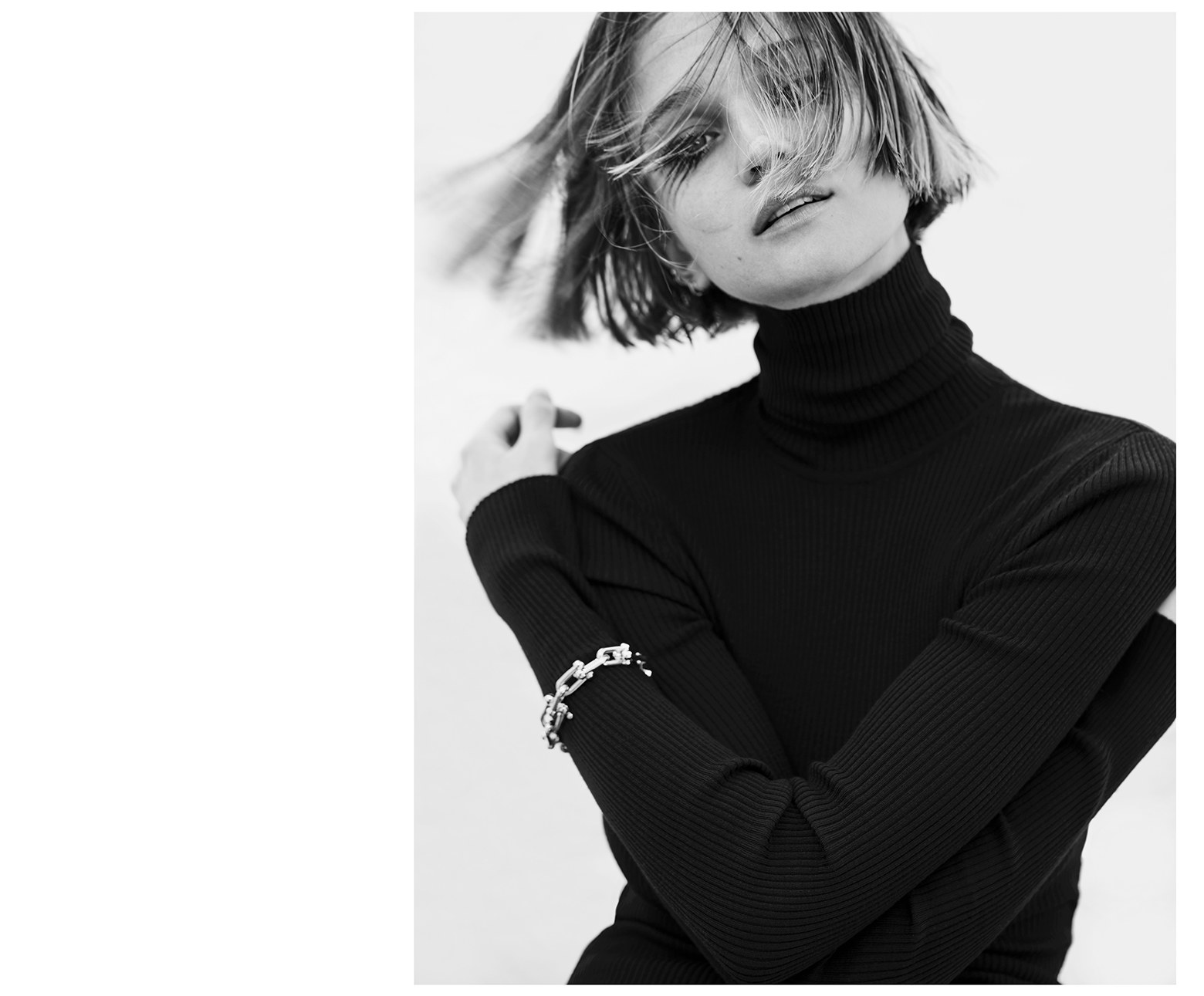 Black and white portrait of a woman with short hair wearing a black turtleneck and silver chain bracelet against a white background.