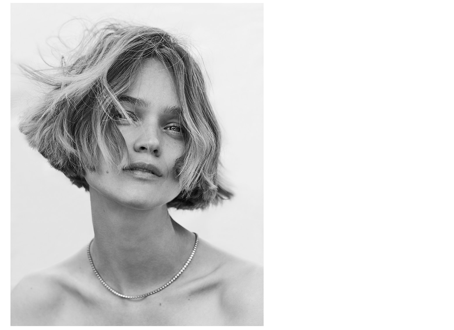 Black and white portrait of a young adult woman with a bob haircut, wearing a delicate necklace, against a bright, neutral background.
