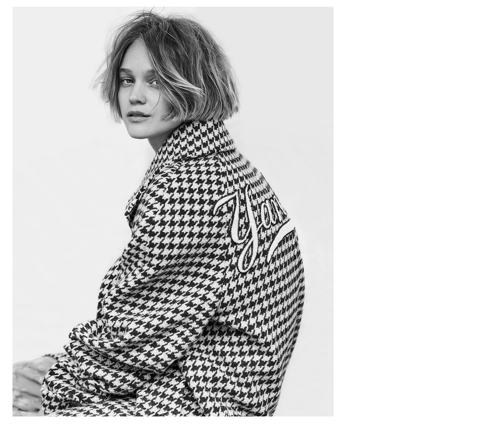 Portrait of a young adult woman with short wavy hair wearing a black and white patterned jacket against a white background in a studio setting.