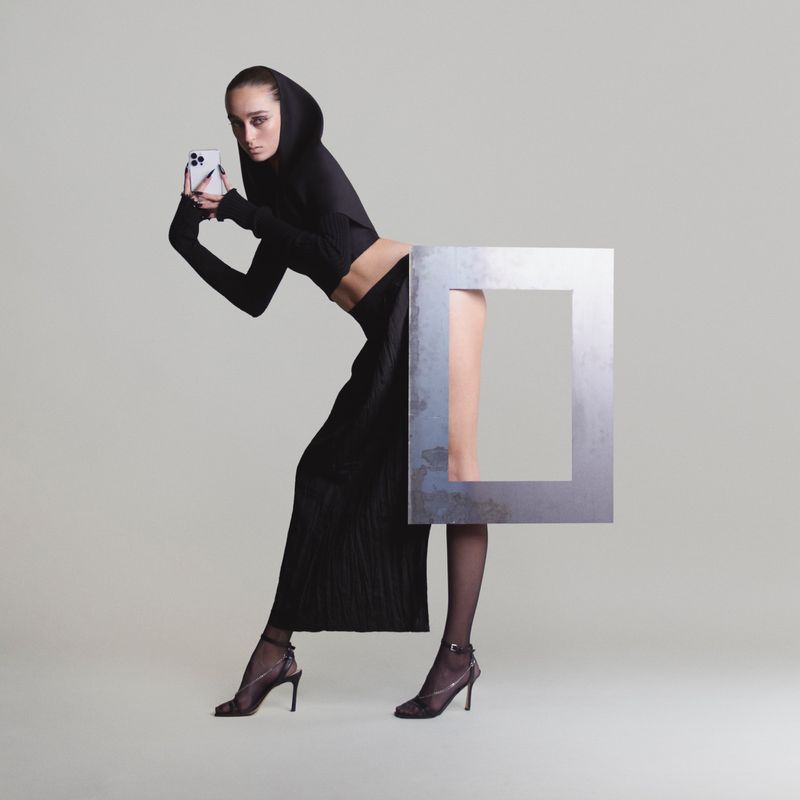 A fashion photo features a slender woman in black attire holding a smartphone, with a geometric frame partially obscuring her lower body against gray.
